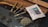 Several rolled cigars or blunts are placed on a wooden surface next to a humidity control packet. There is a display of scattered dried herbs or leaves.