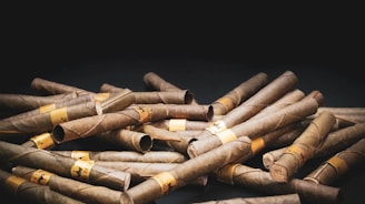A pile of brown rolled cigars is scattered across a dark surface. Each cigar is wrapped in a paper band that has some illegible text. The background is black, emphasizing the texture and color of the cigars.