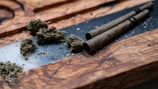 A close-up of loose cannabis buds placed on a wooden surface with two rolled cigars or joints lying beside them. There is some scattered plant material around the objects, and the wooden surface has a rich grain texture.