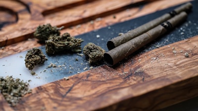 A close-up of loose cannabis buds placed on a wooden surface with two rolled cigars or joints lying beside them. There is some scattered plant material around the objects, and the wooden surface has a rich grain texture.