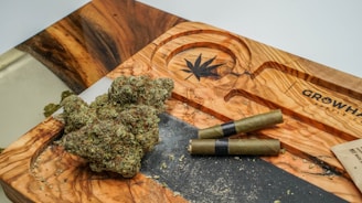 A wooden tray with a carved cannabis leaf design, holding several nugs of cannabis and two pre-rolled joints. The tray features natural wood grain patterns and is situated on a flat surface. Some cannabis crumbs are scattered around the tray.