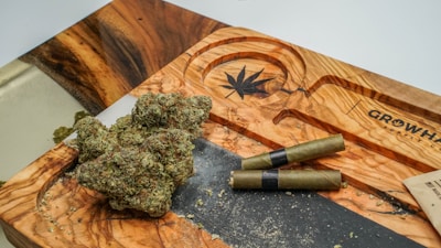 A wooden tray with a carved cannabis leaf design, holding several nugs of cannabis and two pre-rolled joints. The tray features natural wood grain patterns and is situated on a flat surface. Some cannabis crumbs are scattered around the tray.
