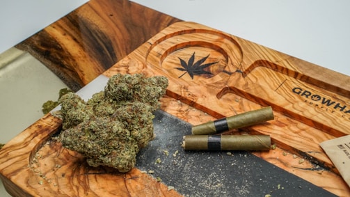 A wooden tray with a carved cannabis leaf design, holding several nugs of cannabis and two pre-rolled joints. The tray features natural wood grain patterns and is situated on a flat surface. Some cannabis crumbs are scattered around the tray.