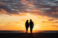 Two people walking hand in hand along a beach at sunset, their silhouettes glowing.
