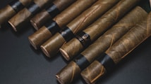 A collection of neatly arranged cigars lies on a dark surface. Each cigar is wrapped in brown leaf with visible veins and features a black band around its center.