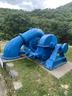 A large industrial machine painted blue sits outdoors on a patch of green grass. It features multiple pipes and metallic parts, including a prominent curved section. The background consists of lush green trees and a cloudy sky.
