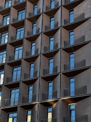 A modern building facade with a grid-like pattern of windows and balconies. Each unit features a dark metallic frame, with reflective glass windows and sleek black railings. The repeating geometric pattern creates a uniform and symmetrical appearance.