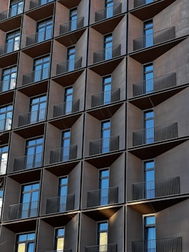 A modern building facade with a grid-like pattern of windows and balconies. Each unit features a dark metallic frame, with reflective glass windows and sleek black railings. The repeating geometric pattern creates a uniform and symmetrical appearance.