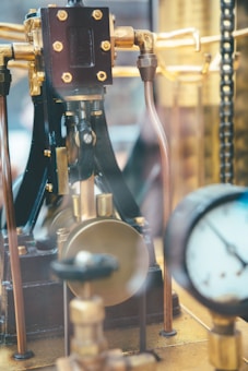 An intricate piece of machinery with metallic components, brass pipes, and a pressure gauge. The mechanisms appear well-crafted, with bolts and joints visibly connected. The background is slightly blurred, emphasizing the central engineering elements.