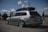 A silver Volkswagen Golf estate car is parked in an open area under a partly cloudy sky. The car is equipped with a roof box and has shiny, modified rims. Several people are in the background, some standing near another car with an open hood.