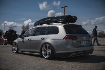 A silver Volkswagen Golf estate car is parked in an open area under a partly cloudy sky. The car is equipped with a roof box and has shiny, modified rims. Several people are in the background, some standing near another car with an open hood.