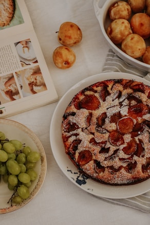 A table setting featuring a freshly baked fruit tart with powdered sugar, an open cookbook displaying recipes, a cluster of green grapes on a decorative plate, and a bowl filled with round fruits. The scene evokes a cozy and homey atmosphere with a focus on baking and fruit.