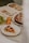 A table setting with a slice of fruit tart on a plate, a whole tart dusted with powdered sugar on a dish, an open cookbook with images of food, and a cluster of green grapes next to two yellow apples.