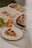 A table setting with a slice of fruit tart on a plate, a whole tart dusted with powdered sugar on a dish, an open cookbook with images of food, and a cluster of green grapes next to two yellow apples.