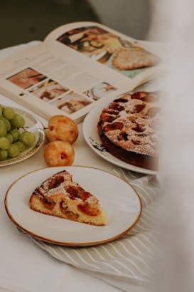 A table setting with a slice of fruit tart on a plate, a whole tart dusted with powdered sugar on a dish, an open cookbook with images of food, and a cluster of green grapes next to two yellow apples.