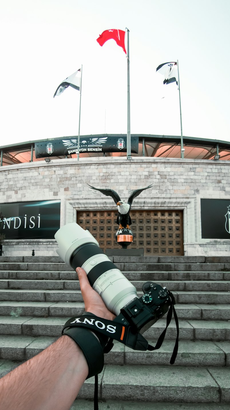 A hand holding a camera with a long white telephoto lens up in front of a stadium entrance with flags and an eagle statue
