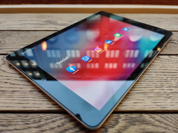 A tablet is placed on a wooden table, displaying a screen with various social media app icons such as Facebook, Instagram, LinkedIn, and Twitter. The reflection of lights above adds a dynamic visual effect to the screen.