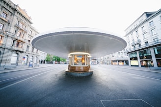 A modern, circular tram or bus stop structure sits at the center of an empty city street, framed by historic European-style buildings on either side. The wide canopy provides shelter, with benches and advertisements visible inside. The area is well-lit and appears clean, with no people present.