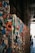 A wall covered in a dense layer of colorful chewing gum, featuring various shapes and hues. The gum is applied haphazardly, creating a textured, three-dimensional surface. The background shows a brick wall and an industrial setting with pipes, giving the scene a gritty urban atmosphere.