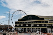 A large building with the words 'Seattle Aquarium' prominently displayed on the facade. In the background, a Ferris wheel rises majestically against a partly cloudy sky. The scene is bustling with people and vehicles, while orange traffic cones and barriers are visible on the street.