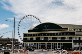 A large building with the words 'Seattle Aquarium' prominently displayed on the facade. In the background, a Ferris wheel rises majestically against a partly cloudy sky. The scene is bustling with people and vehicles, while orange traffic cones and barriers are visible on the street.