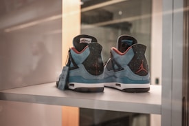 A pair of blue and black sneakers with red accents is displayed on a shelf. The sneakers feature a distinct design with a visible tag hanging from one shoe. The iconic Jumpman logo is visible on the back of the heels.