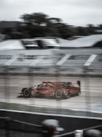 A red racing car speeds along a racetrack, slightly blurred to emphasize motion. The scene is viewed through protective fencing, with spectators visible at the bottom edge. Gray skies and track infrastructure are seen in the background, contributing to a dynamic and intense atmosphere.