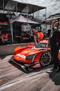 A bright red race car with various sponsor logos is positioned in the pit area. People around the vehicle are wearing racing suits and headsets, indicative of a race event setting. Behind the car, there are tents and platforms filled with spectators. The car is sleek with a modern design and racing tires.
