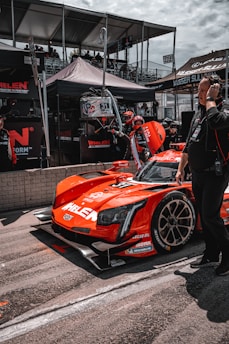 A bright red race car with various sponsor logos is positioned in the pit area. People around the vehicle are wearing racing suits and headsets, indicative of a race event setting. Behind the car, there are tents and platforms filled with spectators. The car is sleek with a modern design and racing tires.