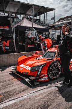 A bright red race car with various sponsor logos is positioned in the pit area. People around the vehicle are wearing racing suits and headsets, indicative of a race event setting. Behind the car, there are tents and platforms filled with spectators. The car is sleek with a modern design and racing tires.