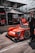 A bright red race car with various sponsor logos is positioned in the pit area. People around the vehicle are wearing racing suits and headsets, indicative of a race event setting. Behind the car, there are tents and platforms filled with spectators. The car is sleek with a modern design and racing tires.