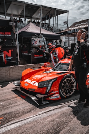 A bright red race car with various sponsor logos is positioned in the pit area. People around the vehicle are wearing racing suits and headsets, indicative of a race event setting. Behind the car, there are tents and platforms filled with spectators. The car is sleek with a modern design and racing tires.