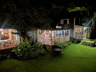 A charming, rustic house with a wooden porch is warmly lit against the night, surrounded by lush greenery. The structure features a sloped roof and large windows, creating a cozy atmosphere. A large pot with aquatic plants sits on the well-manicured lawn, and a set of old stone steps leads to the porch.