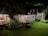 A charming, rustic house with a wooden porch is warmly lit against the night, surrounded by lush greenery. The structure features a sloped roof and large windows, creating a cozy atmosphere. A large pot with aquatic plants sits on the well-manicured lawn, and a set of old stone steps leads to the porch.