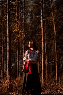 A woman in traditional attire stands in a forest, basking in dappled sunlight. She wears a white embroidered blouse with red beads and a red sash, accompanied by a long dark skirt. Her expression is serene and contemplative as she gazes into the distance.