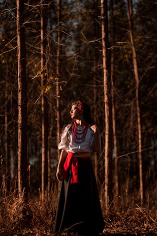 A woman in traditional attire stands in a forest, basking in dappled sunlight. She wears a white embroidered blouse with red beads and a red sash, accompanied by a long dark skirt. Her expression is serene and contemplative as she gazes into the distance.