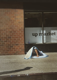 A person is sitting on a sidewalk against a brick wall, partially covered by a blanket. A storefront window with the words 'up market' is partly visible. There is a bottle placed on the ground next to the person.