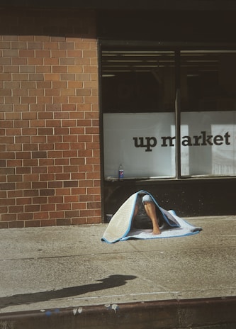 A person is sitting on a sidewalk against a brick wall, partially covered by a blanket. A storefront window with the words 'up market' is partly visible. There is a bottle placed on the ground next to the person.