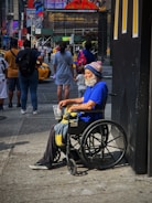 An older man with a white beard is sitting in a wheelchair on a busy city sidewalk. He is wearing a bright blue shirt and a winter hat with the word 'GIANTS' visible. He holds a plastic cup and a blanket rests on his lap. Several people are walking by in the background, along with yellow taxis. There are large advertisements and neon signs on nearby buildings, giving the scene a typical urban atmosphere.