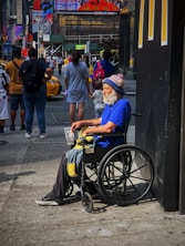 An older man with a white beard is sitting in a wheelchair on a busy city sidewalk. He is wearing a bright blue shirt and a winter hat with the word 'GIANTS' visible. He holds a plastic cup and a blanket rests on his lap. Several people are walking by in the background, along with yellow taxis. There are large advertisements and neon signs on nearby buildings, giving the scene a typical urban atmosphere.