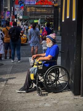 An older man with a white beard is sitting in a wheelchair on a busy city sidewalk. He is wearing a bright blue shirt and a winter hat with the word 'GIANTS' visible. He holds a plastic cup and a blanket rests on his lap. Several people are walking by in the background, along with yellow taxis. There are large advertisements and neon signs on nearby buildings, giving the scene a typical urban atmosphere.