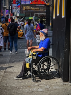 An older man with a white beard is sitting in a wheelchair on a busy city sidewalk. He is wearing a bright blue shirt and a winter hat with the word 'GIANTS' visible. He holds a plastic cup and a blanket rests on his lap. Several people are walking by in the background, along with yellow taxis. There are large advertisements and neon signs on nearby buildings, giving the scene a typical urban atmosphere.