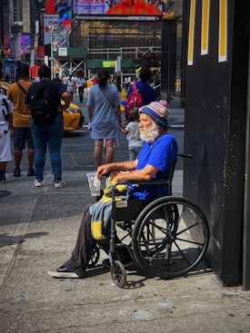 An older man with a white beard is sitting in a wheelchair on a busy city sidewalk. He is wearing a bright blue shirt and a winter hat with the word 'GIANTS' visible. He holds a plastic cup and a blanket rests on his lap. Several people are walking by in the background, along with yellow taxis. There are large advertisements and neon signs on nearby buildings, giving the scene a typical urban atmosphere.