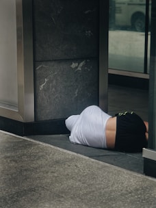 A person lies on the ground in a partially covered area, wearing a white shirt and black shorts. The location appears to be an urban setting, possibly near a building entrance, characterized by dark stone tiles and reflective glass surfaces.