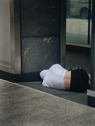 A person lies on the ground in a partially covered area, wearing a white shirt and black shorts. The location appears to be an urban setting, possibly near a building entrance, characterized by dark stone tiles and reflective glass surfaces.