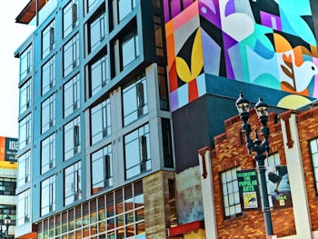 A city scene featuring modern architecture alongside an older brick building. The modern building has large glass windows reflecting the surroundings, while the brick building displays posters that suggest it might be a cultural or entertainment venue. A prominent mural with geometric shapes and a stylized bird adds color and vibrancy to the scene. A classic street lamp stands in the foreground, contributing to the urban ambiance.