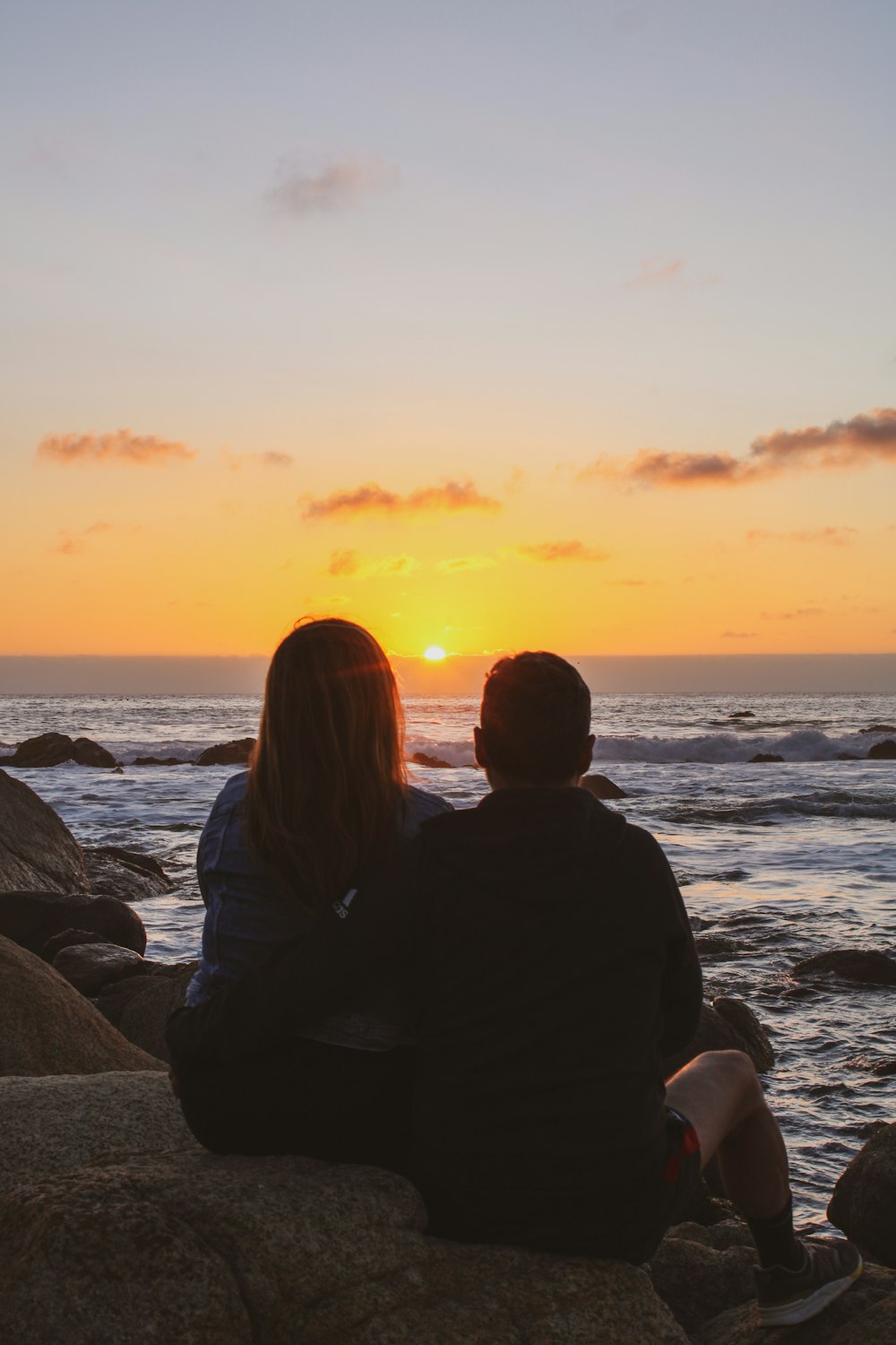 a man and woman sitting on a rock looking at the ocean