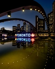 A night view of the Toronto city sign and skyline, featuring illuminated buildings reflected in a calm body of water. The large iconic 'TORONTO' sign is brightly lit, incorporating a red maple leaf symbol. The structures around are adorned with light archways, adding to the urban aesthetic.