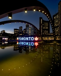 A night view of the Toronto city sign and skyline, featuring illuminated buildings reflected in a calm body of water. The large iconic 'TORONTO' sign is brightly lit, incorporating a red maple leaf symbol. The structures around are adorned with light archways, adding to the urban aesthetic.