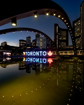 A night view of the Toronto city sign and skyline, featuring illuminated buildings reflected in a calm body of water. The large iconic 'TORONTO' sign is brightly lit, incorporating a red maple leaf symbol. The structures around are adorned with light archways, adding to the urban aesthetic.
